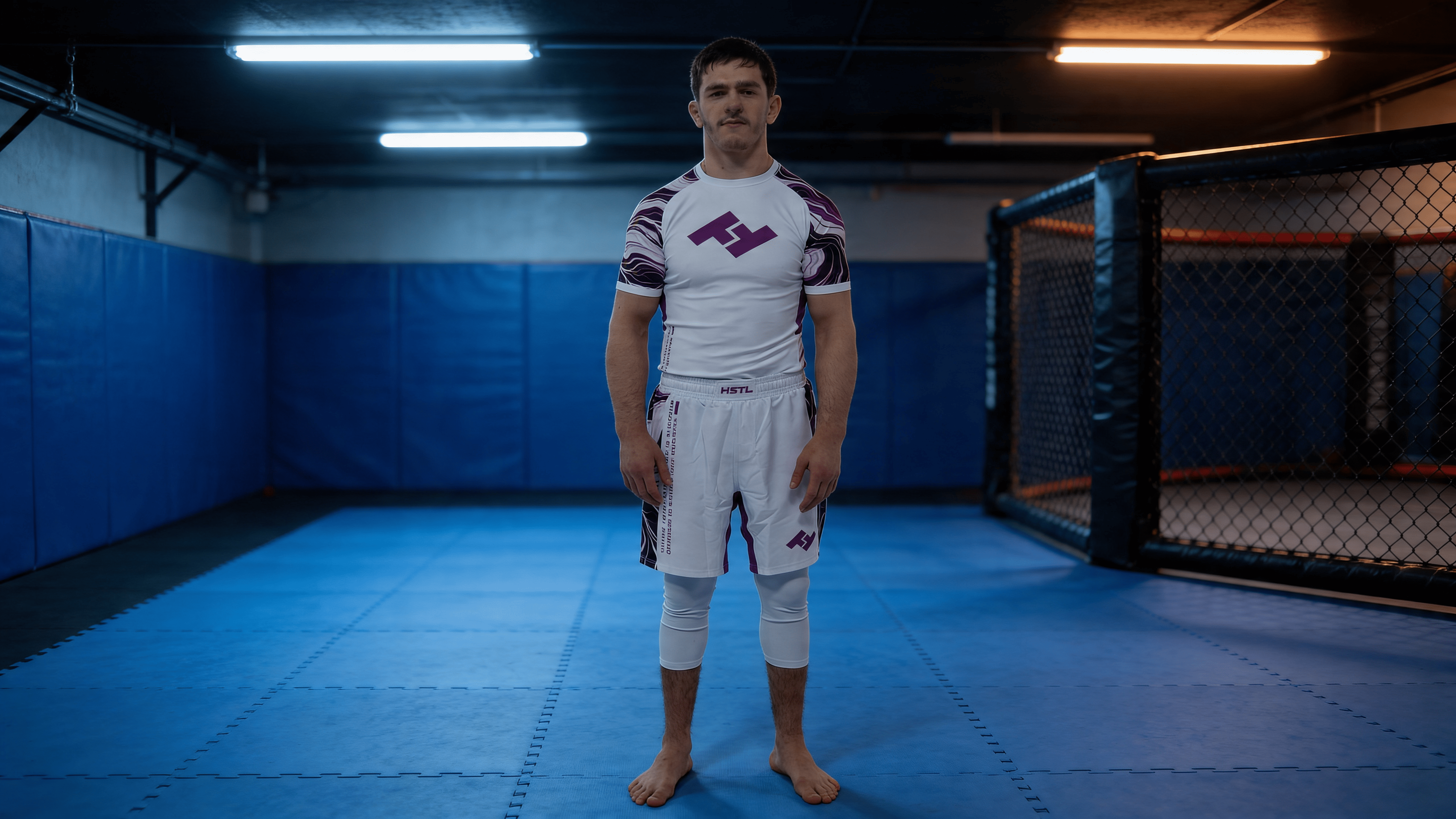 Combattant portant un rashguard Hustle Fightwear sur tatamis bleus dans une salle de grappling avec cage MMA