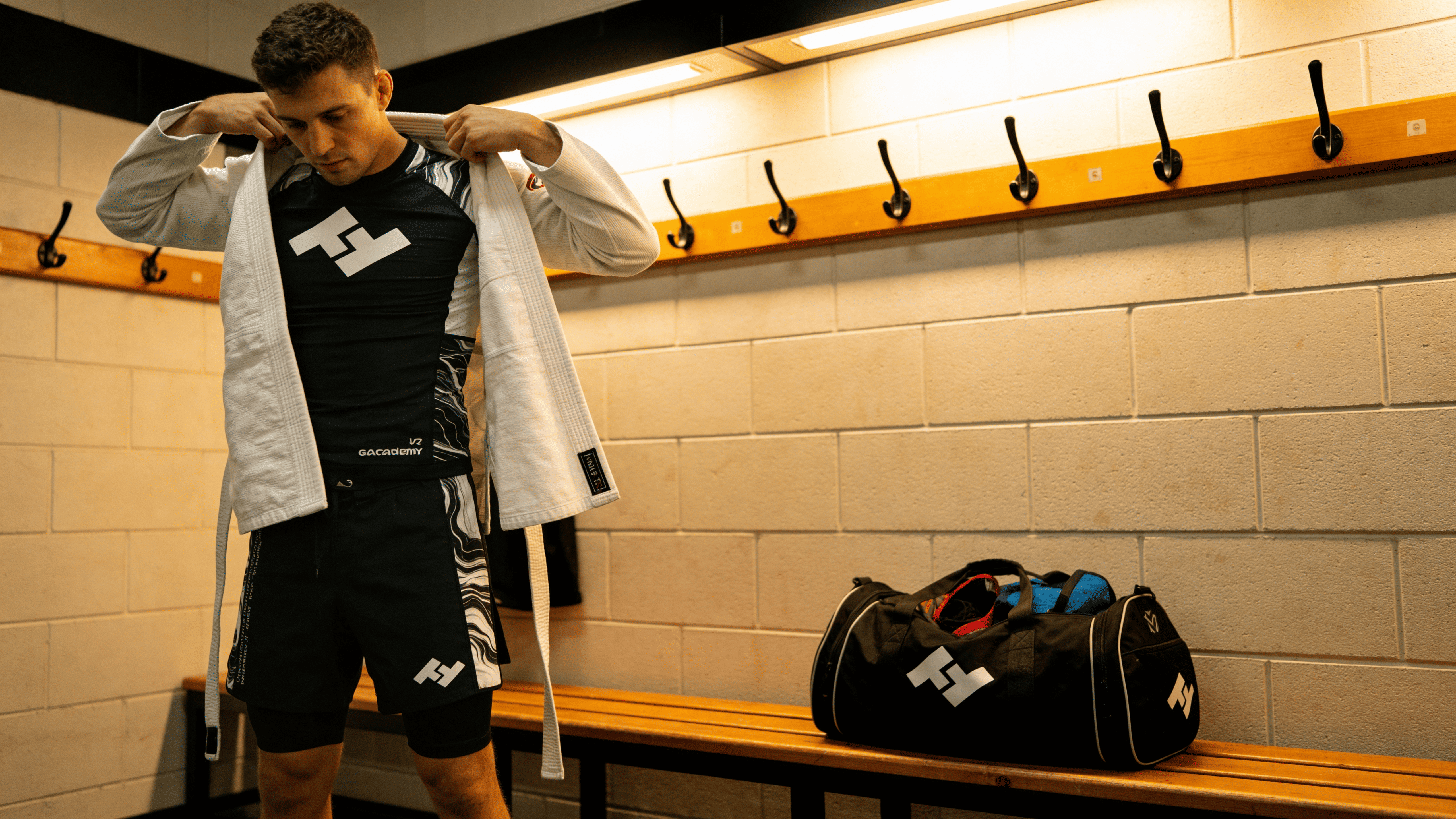 Pratiquant JJB enfilant son kimono par-dessus un rashguard Hustle Fightwear dans un vestiaire